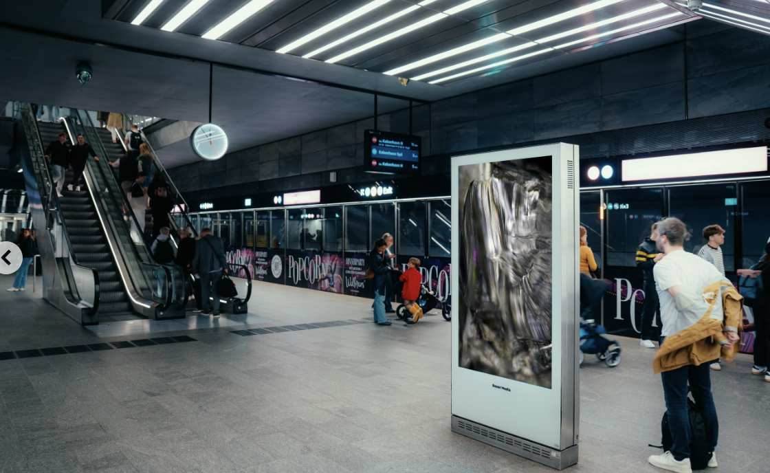 Digital DOOH-skærm i metroens stationshal ved rulletrapper og afgangstavler.