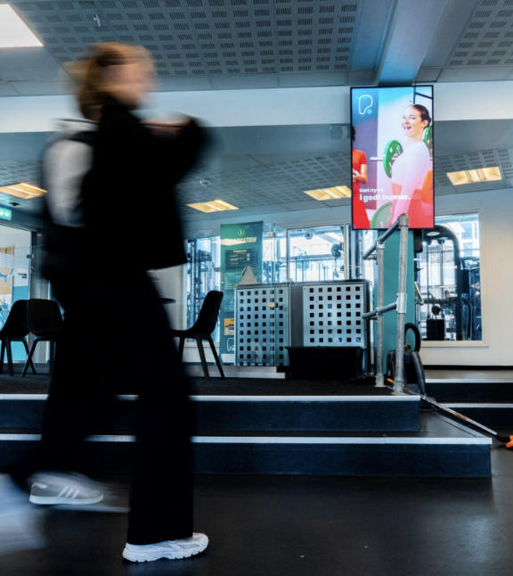Digital annonceskærm på en søjle i et PureGym-center med en person, der går forbi i træningstøj.